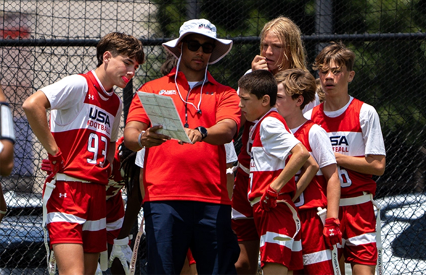 USA Football coach reviewing playbook with athletes during Select Bowl team huddle.
